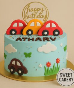 Car theme birthday cake by Sweet Cakery in Bangalore featuring fondant cars, clouds, grass, flowers, and a golden topper on soft blue cream icing.