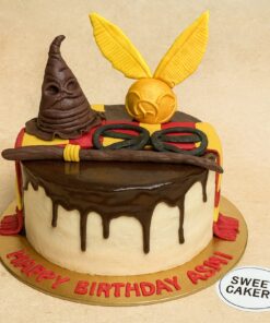 Harry Potter birthday cake by Sweet Cakery featuring cream finish, Sorting Hat, Golden Snitch, wand, glasses, and Gryffindor scarf on top