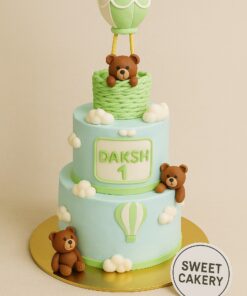 Two-tier pastel cream cake with hot air balloon theme, teddy bears, and clouds. Created by Sweet Cakery with custom decorations and realistic styling.