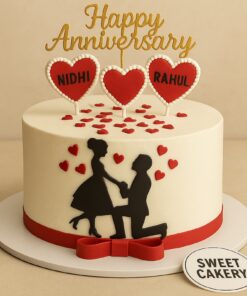 A romantic anniversary cake with black couple silhouette, red heart toppers with names, golden “Happy Anniversary” topper