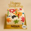 Square cream birthday cake with fondant crayons, scissors, ruler, buttons, and colour splashes in arts and crafts theme.
