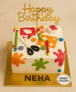 Square cream birthday cake with fondant crayons, scissors, ruler, buttons, and colour splashes in arts and crafts theme.