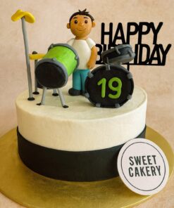A professional photo of a Drum Theme Birthday Cake by Sweet Cakery in Bangalore, featuring a fondant drummer figurine with a green drum set and band-style decorations.