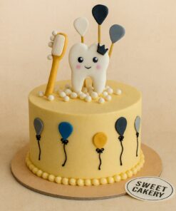 A yellow cream cake with a smiling tooth topper, toothbrush, balloons, and fondant details, from Sweet Cakery Bangalore, designed for a baby’s first tooth celebration.